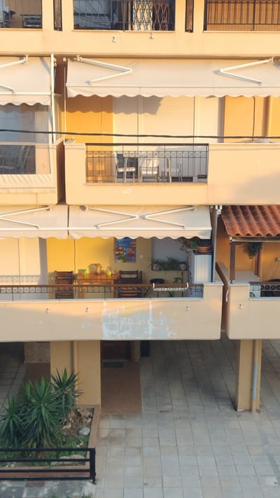 The image shows a multi-story building with balconies and awnings. The building has light-colored walls, possibly beige or cream, and some areas show peeling paint. The awnings are white and appear to be made of fabric, supported by metal frames. Some balconies have metal railings, and there are windows with security grilles on some floors.

On one balcony, there are plants in pots, suggesting an outdoor space for relaxation or decoration. There is also a colorful painting visible on the wall of one of the balconies, adding a decorative element. The scene appears to be an exterior view of an apartment building or similar structure.

The lighting suggests it might be late afternoon or early morning, with the sun casting shadows. The location appears to be in a residential area, possibly a town or city. There is no clear indication of any specific activity or event taking place, other than the general appearance of a building.

The image is oriented such that the building runs vertically, suggesting the photo may have been taken looking upwards or the camera was rotated. The ground outside the building is paved with square tiles. In the foreground, on the right side, there is some greenery, possibly a small garden or planted area with spiky plants. A black metal fence or railing is also visible on the right edge of the image.

The overall impression is a somewhat weathered but functional building, likely in a Mediterranean setting given the architecture and outdoor elements. There is no visible text in the image.