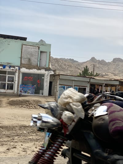 The image depicts a daytime street scene, likely in a rural or semi-urban area of Dand, Afghanistan. In the immediate foreground, parts of a motorcycle are visible, including handlebars, chrome headlights, and a red spring shock absorber, suggesting the photo was taken from a vehicle. Plastic bags are tied to the motorcycle's front, and a purple patterned cloth covers another part.
Beyond the motorcycle, a dirt road leads past several buildings. On the left, a two-story building with a light mint green facade and peeling paint stands out. Its second story features a boarded-up window and a white door. Solar panels are visible on its roof. The ground in front of this building shows piles of bricks. Below, shopfronts with glass windows and doors display signs.
Further down the street, more single-story shops with corrugated metal roofs are visible. In the mid-ground, a utility pole and a small green tree break the view of the vast, rugged, and arid mountains that dominate the background under a partly cloudy, bright sky.
Visible text on the shopfronts includes "انورالدین در ملتون" (Anwaruddin in Melton - likely a business name or location), contact numbers "0706666953" and "0705830758," and "دروازه" (Door/Gate). Other Farsi text such as "هنمایی معاملات" (Guidance/Help in Transactions) and "تماس شميري" (Contact Number) are also present. The scene captures daily life in an area with a rugged natural landscape.