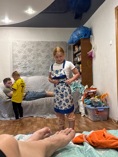 The image shows a family in an indoor living space. In the foreground, a person's legs and feet are visible, lying on a turquoise blanket. Further back, a young girl with blonde hair is standing and wearing a white t-shirt and a blue and white floral apron. To her left, a young boy in a yellow t-shirt with "RONALDO 7" printed on it is facing a man lying on a textured grey couch. The man is wearing a green t-shirt and blue jeans. The room has wooden flooring and white walls. A dark grey ceiling with recessed lighting is visible. A large mattress with a floral pattern leans against the wall behind the couch. To the right, a wooden shelf unit is filled with various items, including blue plastic bags and other assorted objects. In front of the shelves, a clear plastic storage bin filled with toys and other items is on the floor. A bright orange towel is also visible on the turquoise blanket in the foreground. The overall impression is a casual, domestic scene. The image is identified as being taken in the city of Valday, Russia.