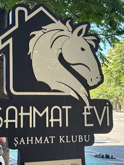 A black sign with a white outline of a house and a white silhouette of a horse's head is mounted on a pole. Below the horse's head, in large white letters, are the words "ŞAHMAT EVİ". Below that, in smaller white letters, are the words "ŞAHMAT KLUBU". The sign is outdoors, with green foliage and blue sky visible in the background. The sign appears to be for a chess club, as "Şahmat" is the Azerbaijani word for chess. The sign is likely located in Baku, Azerbaijan, given the Azerbaijani language on the sign. The area around the sign appears to be a street, with a road and sidewalk visible.