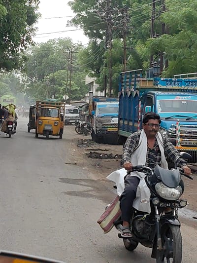 In Jambhuldhaba, India, a man wearing glasses and a checkered shirt with a white scarf draped around his neck rides a black motorcycle on a paved road. He is seen from the chest down, with his hands on the handlebars. A white piece of fabric is draped over the motorcycle's seat and extends to his left side, alongside a bag with red and yellow stripes. To the right of the motorcycle, a blue truck with "GOODS CARRY" written on it is partially visible. Behind the motorcycle, an orange auto-rickshaw is stopped. Further in the background, other vehicles, including a green bus, are present. The environment appears to be a street with trees and utility poles lining the side of the road. The weather seems overcast, and the road surface is wet in places, suggesting recent rain or damp conditions.