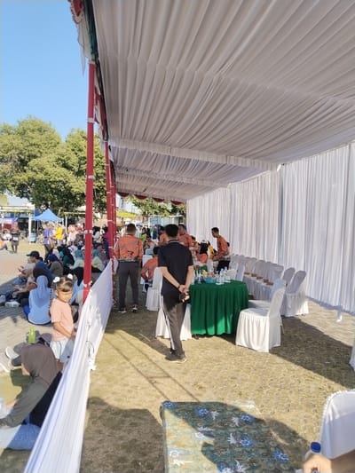 This image captures an outdoor event under a large tent structure in Sragen, Indonesia, on a clear, sunny day. The tent features a white, horizontally draped fabric ceiling and white pleated side curtains, supported by several red vertical poles.
On the left side of the frame, outside the immediate tent area, a crowd of people is gathered, predominantly seated on the ground. Many individuals, especially women, are wearing head coverings (hijabs). A young girl with pigtails, dressed in a light pink shirt and light blue shorts, stands near a white fabric divider, observing the activity. In the background on the left, green trees are visible, along with glimpses of other small tents and structures.
Beneath the main tent on the right, the ground is a mix of dry grass and paved tiles. A round table covered with a dark green tablecloth holds an assortment of bottled water, drinks, and snacks. Several white chairs with white fabric covers are arranged around this table and extend in a row to the right, some appearing unoccupied. Individuals are standing and moving within this area; a man in a black t-shirt and track pants stands with his back to the camera near the green-covered table, while another man in an orange-patterned shirt (possibly batik) is visible in the mid-ground. In the bottom right foreground, a corner of another table covered with a blue and brown patterned tablecloth is visible. The bright sunlight casts distinct shadows across the ground.
