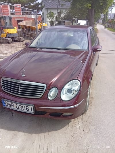 In Wilczyce, Poland, a maroon Mercedes-Benz E-Class W211 sedan is parked on a dirt road, likely near a construction site. The car is facing slightly towards the viewer's left, and its license plate reads "DWR 0308 J". In the background, construction fencing encloses a building under renovation, with a yellow excavator and a yellow dumper truck visible. Beyond the construction, a street lined with trees and houses stretches into the distance, where a person is walking. The sky is overcast. The timestamp "09.06.2026 10:00" and "POCO C75" are superimposed on the lower part of the image.