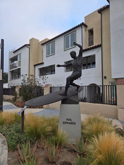 The image shows a bronze statue of a surfer named Phil Edwards in Dana Point, California, USA. The statue depicts Edwards in mid-ride on a surfboard, with one arm raised. The sculpture stands on a concrete pedestal inscribed with "PHIL EDWARDS." It is situated in front of a modern building with a Mediterranean architectural style, featuring stucco exteriors, red tile roofs, and balconies. The surrounding landscaping includes drought-tolerant plants such as ornamental grasses and succulents. A paved pathway with a diamond pattern is visible in the foreground, leading towards the statue. The sky is overcast.