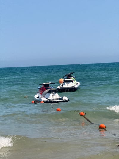 This media file features a high-angle, rotated view of a beach scene. The primary subjects are two jet skis, one white with green accents and the other white and red, floating in the water near the shore. Several orange buoys are visible, some attached to a rope.
The scene is set in a body of water with gentle waves breaking on a sandy shore. The water is a clear, greenish-blue, and the sky above is a pale, cloudless blue. The lighting suggests it is daytime, likely a sunny day given the brightness and clear sky. The presence of jet skis and buoys indicates a recreational area, possibly a designated water sports zone.
One jet ski appears to have a person on it, wearing a life vest, suggesting activity. The other jet ski is also in the water. The overall atmosphere is one of a pleasant day at the beach, with potential for water activities. No visible text is present in the image.