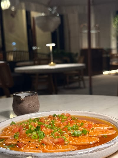 The image shows a plate of sashimi with chopped tomatoes and green onions on a white table. Next to the plate is a small, brown ceramic container with a white rim. In the background, out of focus, is an interior scene with tables and chairs, presumably a restaurant or dining area. The location is دبي, United Arab Emirates.