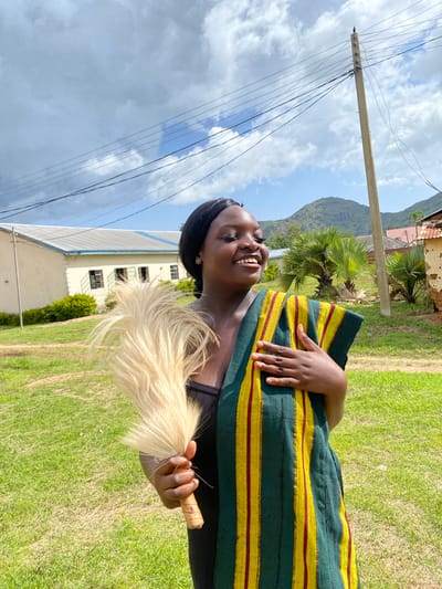 This is a portrait of a young Black woman outdoors. She is wearing a traditional garment with thick horizontal stripes of dark green, yellow, and thin red. Her left hand rests on her chest, covering a portion of the garment, and her right hand holds a light-colored whisk or fly-swatter made of animal hair. The woman is smiling broadly, with her head tilted slightly back and her eyes looking upwards. She has dark braided hair pulled back.
The setting appears to be rural or semi-rural. Behind her, there is a well-maintained grassy area. In the mid-ground, a two-story building with a white exterior and a light-colored roof is visible, along with some green shrubbery. Further in the background, there are palm trees and a lush, green mountainside under a cloudy sky. A utility pole with numerous wires extends from the left side of the frame towards the upper left corner.
The lighting suggests it is daytime, likely afternoon, given the bright sunlight illuminating the grass and the building. The weather appears partly cloudy, with patches of blue sky visible between the clouds. The overall mood of the image is joyful and relaxed, primarily conveyed by the woman's happy expression. The location context is Wang, Nigeria. There is no visible text in the image.