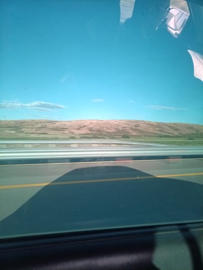 The media file is a photograph taken from inside a vehicle, looking out. The foreground shows the interior of the car, including a shadow cast on the dashboard and a portion of the car's frame. The view through the windshield reveals a road with a yellow line. Beyond the road, there is a grassy field and a rolling, barren hill. The sky is a clear, bright blue with a few wispy clouds. The scene suggests daytime with good weather. Reflections on the windshield indicate the presence of the sun and potentially other vehicles or structures. The landscape appears to be semi-arid or steppe-like, consistent with the location context of Субхоз, Kazakhstan. No people are clearly visible, but the presence of the vehicle implies human activity. The overall impression is one of travel through a rural or undeveloped area under a wide-open sky.