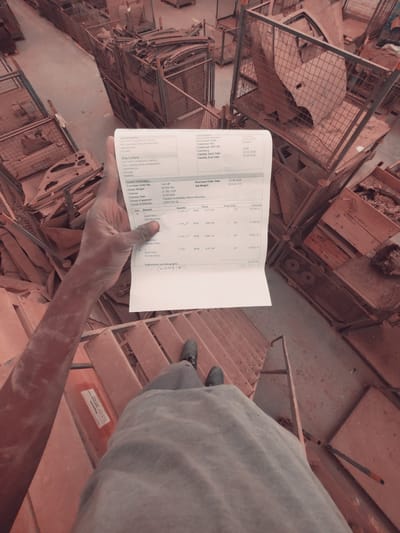 The image depicts a person's hands holding a document that appears to be an invoice or receipt, positioned in front of a backdrop of industrial scaffolding and metal cages filled with scrap metal. The document displays various details including "Sold To-Party," "Ship To-Party," "Header Information," and itemized entries with "Item," "Material," "Quantity," "Price," and "Amount." Specific text visible on the document includes "CRAFT WORKSHOP LIMITED," "KADUNA-ZARIA EXPRESSWAY," "UNGUWAN KAJI," "KADUNA," "NIGERIA," "Purchase Order Date 23.06.2026," "Gross Weight 23.024 KG," and item descriptions such as "HUB BEARING." The environment suggests an industrial setting, possibly a scrap yard or manufacturing facility, with stacks of metal components and structures. The lighting is somewhat dim, and the overall tone of the image is utilitarian. The location, Kakuri Gwari, Nigeria, is inferred from the text on the document and the context of the industrial setting.