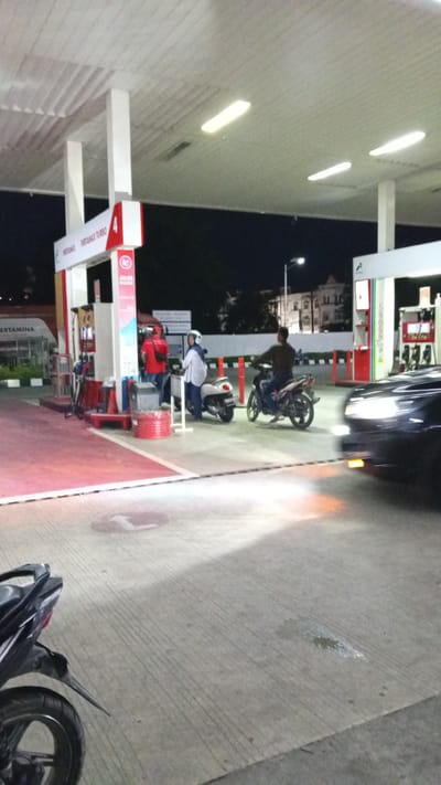 The image depicts a night scene at a Pertamina gas station in Cotbadatunong, Indonesia. The gas station features a white canopy supported by white pillars, illuminated by fluorescent lights. Several people and vehicles are present.
In the foreground, a black motorcycle is parked on the right side of the image, with its rear wheel and fender clearly visible. Further into the scene, on the left, a man in a red shirt stands near a gas pump. To his right, a woman wearing a white hijab and a patterned shirt is seated on a white scooter, interacting with another individual. A man is riding a dark-colored motorcycle, heading away from the camera. To the right of the frame, a dark-colored car is partially visible, its headlights illuminated. The ground is paved concrete, with red markings indicating lanes or parking areas. In the background, buildings and streetlights are visible, suggesting an urban or semi-urban environment. The overall impression is one of a typical night at a busy gas station.