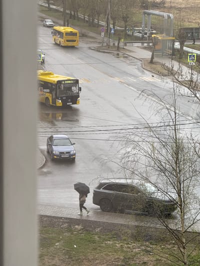 An overhead shot reveals a rainy street scene in what appears to be Ново-Никольское, Russia. Several vehicles are visible: a yellow bus, a smaller silver-blue car, and a dark-colored sedan parked on the sidewalk. A pedestrian with a black umbrella walks on the sidewalk. Overhead power lines traverse the street. A cluster of industrial-looking pipes and a walkway are seen in the background, along with several trees without leaves. The sidewalk is bricked or paved, with grass growing on the edge. The street is wet and reflective, indicating recent rainfall.