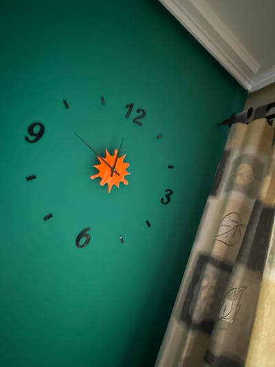 This media file shows a close-up view of a clock with black Arabic numerals on a teal green wall. The clock has an orange, abstract-shaped minute hand and a thin black hour hand. The numerals are scattered around the clock face, with the "12" at the top left, "3" at the top right, and "6" at the bottom right. The scene appears to be indoors, possibly in a home or office. The lighting is somewhat dim, casting subtle shadows. The background features a blurred curtain with a pattern of leaves and dark rectangles. There is also a white molding detail visible in the top left corner. The image is likely a photograph or a high-resolution screenshot. No people are visible in the frame, and no text is present other than the clock numerals. The overall impression is one of a decorative wall clock in a modern setting.