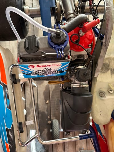 This is a close-up, overhead shot of a racing go-kart engine assembly, likely taken in an outdoor or garage setting, possibly in Sukhaya, Russia. The focus is on the engine components, including a fuel tank with branding that reads "БИЯРЕЛЬ ПЛЮС" and "IUSUPOL TEAM". A clear plastic hose is visible, curving from the top left corner towards the engine, with "New-Line" printed on it. The engine features red and blue parts, a black ribbed hose, and a red spark plug cap. A black rectangular reservoir is mounted below the engine. The go-kart bodywork, featuring blue and white graphics with orange accents, is partially visible on the left side. The background consists of wooden planks, suggesting a workshop or track environment. There are no people or animals visible in the image. The lighting appears to be natural, possibly from an overcast sky or ambient light in a workshop.
