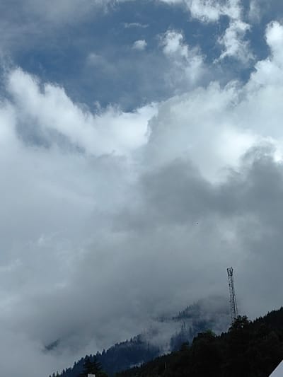 This is a photograph taken from the city of Siyal, India, capturing a mountainous landscape under a cloudy sky. The upper portion of the image is dominated by a dramatic sky filled with both dark and white clouds, with patches of blue sky peeking through. The lower half of the image features a densely forested mountain slope. A tall, slender telecommunications tower stands prominently on the right side of the mountain, its metallic structure contrasting with the natural scenery. The trees on the slope appear dark and green, and some areas of the mountain are obscured by mist or low-hanging clouds, adding a sense of depth and mystery to the scene. The overall atmosphere is overcast and somewhat dramatic, suggesting a possibly cool or damp day.