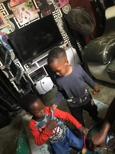 This image depicts an indoor residential scene in Oba-Ile, Nigeria. The background features a wall covered in black and white geometric pattern wallpaper, adorned with multiple framed photographs of individuals and families. A black LG flat-screen television is positioned above a white entertainment stand containing various electronics and cables. To the right of the television, a dusty standing fan is visible. Further right, a dark-colored armchair with significant wear and peeling upholstery is present. Dark fabric drapes with a festive tinsel garland hang on the left. The floor is light-colored and partially covered by mats.
In the foreground, two young Black males are present, along with the visible hands of a third individual. A younger child is positioned kneeling or sitting on the floor, wearing a red long-sleeve shirt over a grey camouflage t-shirt that reads "DAYS OF AWESOME," and blue trousers. This child holds a pink spoon, dipping it into a blue bowl containing a yellowish food substance. Standing behind and slightly to the left of this child is an older boy, dressed in a navy blue long-sleeve shirt with colorful graphics and black trousers. He holds a small, light-brown, irregular-shaped snack in his left hand, and a blue spoon in his right hand, positioned near the shoulder of the kneeling child. On the far right, two dark-skinned hands are visible, interacting with the blue bowl, one appearing to support it while the other's finger points towards the contents. The children appear focused on the food and their interaction.