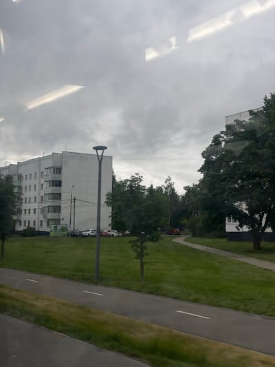 The image depicts a grey, overcast sky seen through a window, with reflections of ceiling lights. Outside, a residential building with multiple stories and balconies is visible, surrounded by trees. A paved path and a grassy area are also present. Several cars are parked along the side of the road near the building. The overall scene suggests a cloudy day in a residential area, likely in Yakovlevskoye, Russia, as indicated by the location context. The image is taken from an interior perspective looking outwards, and the orientation of the image is vertical. No people are visible, and there is no discernible text.