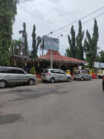 This image captures a street scene in Situbondo, Indonesia, during what appears to be an overcast daytime. The foreground and middle ground are dominated by a paved asphalt road, where several light-colored cars, primarily minivans or small SUVs, are parked parallel to the curb.

On the left side of the frame, a building with a distinctive reddish-brown tiled roof is visible. This structure appears to be a commercial establishment. Above the building, a large rectangular billboard or sign is mounted; however, any text or imagery on it is indistinct and illegible due to blurriness.

Behind and around the building, numerous tall, slender trees with dense green foliage rise prominently against the cloudy sky. Utility poles and overhead electrical wires crisscross the upper portion of the scene. The road surface exhibits some darker patches, possibly wet spots or oil stains. No people are visible, and the scene depicts a quiet moment with parked vehicles, suggesting a typical roadside setting in an urban or suburban area.