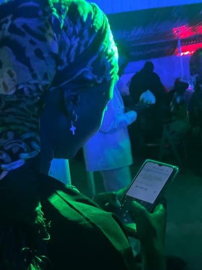 The image shows a close-up of a person's head and hands in a dimly lit environment, possibly a club or party, with green and blue lighting. The person is wearing a patterned headscarf and a cross earring. Their hands are holding and interacting with a smartphone. In the background, other people are visible, some appearing as silhouettes. The scene is characterized by low light conditions and artificial lighting, typical of nighttime social gatherings. No specific landmarks or contextual clues in the image suggest the location is Wumat, Nigeria.