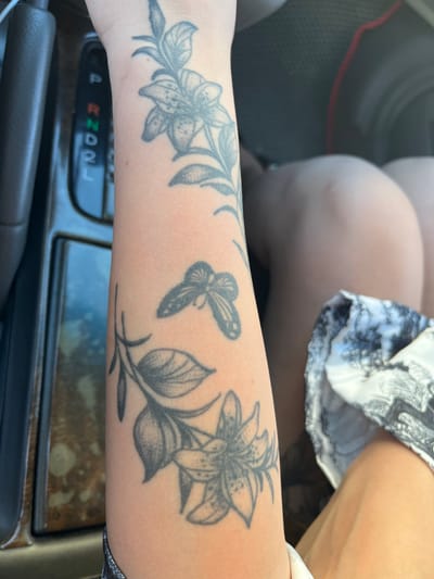 A person's arm with a tattoo of flowers and a butterfly is visible in the foreground, positioned next to a car's gear shift. In the background, a person's legs and knees are visible, with a portion of a patterned skirt. The scene appears to be inside a vehicle. No elements in the image suggest the location of Konopliane, Ukraine.