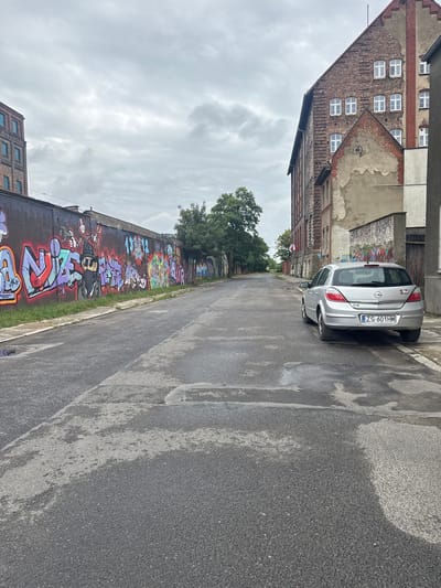 The image depicts a street in Szczecin, Poland, characterized by a row of industrial-style brick buildings adorned with extensive graffiti on the left side of the road. The street itself appears to be asphalt, wet with puddles, suggesting recent rain. A silver hatchback car is parked on the right side of the street, facing away from the camera. The sky is overcast, contributing to the muted color palette of the scene. The overall impression is one of an urban environment, possibly in a less-maintained or industrial district, with a strong presence of street art. The license plate of the car is visible, reading "ZS 601HM".