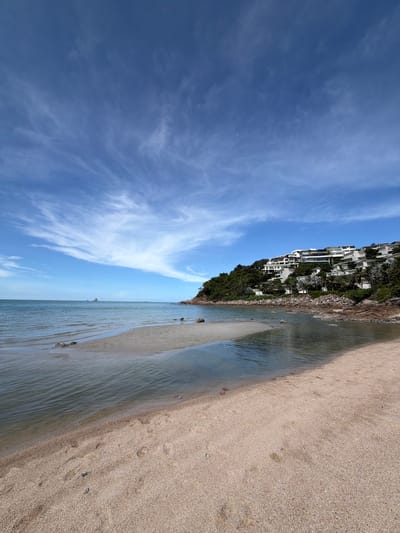 This is a full, vertical shot of a beach and ocean in Choeng Mon, Thailand. The foreground is a sandy beach with some footprints visible. The water is clear and calm, with gentle waves lapping the shore. In the mid-ground, a small sandbar emerges from the water. The background features a lush green hillside dotted with modern white villas, characteristic of the upscale resorts found in the area. The sky is a vibrant blue with wispy white clouds, indicating good weather. A small boat is visible on the horizon in the distance. The scene is peaceful and idyllic, evoking a sense of tropical paradise.