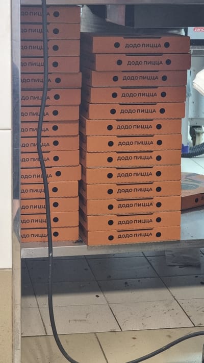 The image shows a stack of orange pizza boxes with the text "ДОДО ПИЦЦА" in Russian, meaning "Dodo Pizza," printed on them. The boxes are stacked in two columns, one to the left and one to the right, on a metal shelf. A black cable runs vertically down the center of the image, obscuring part of the left stack of boxes. The environment appears to be a storage or preparation area, possibly a kitchen or stockroom, with tiled flooring visible below the shelf. A small red and white logo, possibly a brand name, is visible on the far right of the shelf. No people are present in the image. The scene is a straightforward depiction of stacked pizza boxes.
