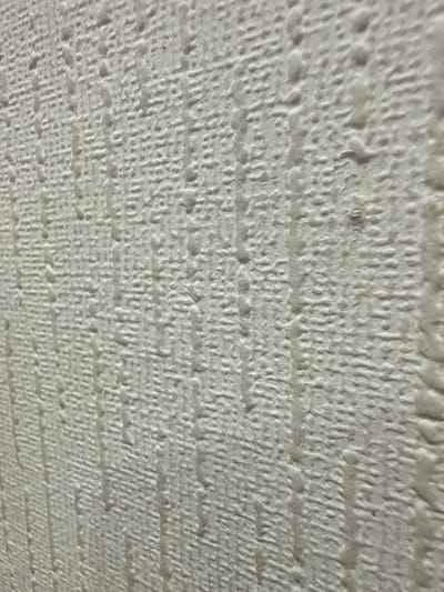 The image displays a close-up, angled view of a textured, off-white wallpaper. The wallpaper has a raised, irregular pattern that appears to be a series of small bumps and ridges. Vertical streaks of slightly darker shade are visible, suggesting a possible moisture stain or a textured line within the wallpaper. The lighting is diffuse, casting soft shadows that emphasize the texture. There are no people, animals, or discernable objects beyond the wallpaper itself. The perspective is from a low angle, looking upwards and to the right. The scene is indoors and the specific location in Angarsk, Russia, cannot be determined from the visual information provided.