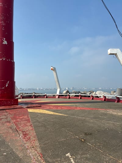 The image shows a concrete pier extending into the sea. Along the edge of the pier are large rocks, with a cityscape visible in the distance. On the right, the pier surface is marked with yellow and red stripes. A red railing runs along the foreground of the pier, and metal support structures are positioned above and at the far end. The sky is blue with some clouds. The location is likely in Coatzacoalcos, Mexico, given the coastal setting and urban landscape. There are no people visible in the scene.