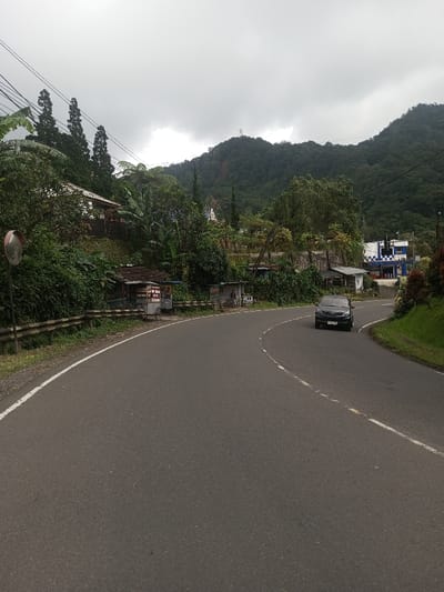 A winding asphalt road stretches through a lush, green landscape under a cloudy sky. Dense vegetation and small structures line the roadside, transitioning into a mountainous backdrop. A dark car approaches, positioned on the right side of the road. The overall impression is a tranquil, somewhat overcast, natural environment intersected by human infrastructure.