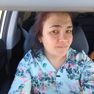 The media file is a close-up, selfie-style photograph of a woman sitting in the passenger seat of a car. The woman has medium-toned skin with freckles and reddish-brown hair. She is wearing a light blue shirt with a floral pattern featuring pink and green leaves and flowers. She is looking towards the camera with a neutral expression, and her head is slightly tilted. A seatbelt is visible across her chest, and the interior of the car, including the headrest and part of the dashboard, is also visible. A small, gold-toned earring with small gems is in her left ear. There is a small metal tag on the front of her shirt with the word "FASHION" written on it in English, backwards and upside down. The lighting suggests it is daytime. The overall scene is casual and personal, likely taken during a car journey. The location context indicates this may be Bulgakovo, Russia.