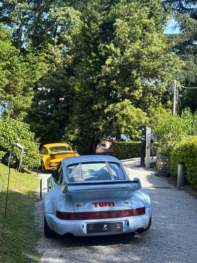 The image depicts the rear of two sports cars, a light blue model with "TUFFI" in red lettering on its rear spoiler, and a yellow car parked behind it. Both are positioned on a gravel driveway beside a grassy area. Lush greenery surrounds the cars, including a large tree that dominates the left side of the frame. In the background, there's a metal structure resembling a security post and further greenery. This scene is within the Lake Como region of Italy.