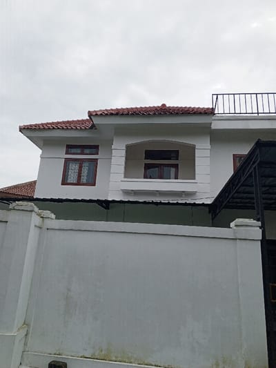 A two-story white building with a red tile roof sits behind a tall, gray concrete wall. The building has multiple windows framed in dark wood, including a central window within an arched balcony. A black metal railing sits atop the second story. Adjacent to the building is a dark, framed structure. The wall shows signs of weathering and moss growth near the base. The sky is overcast.