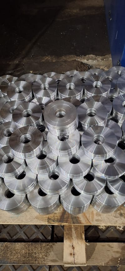 The image shows a large pile of identical, circular metal components with a hole in the center, stacked on a wooden pallet. The components appear to be machined and have a metallic, silver finish with visible machining marks. The background is dark and appears to be a workshop or industrial setting, with a portion of a blue machine visible on the right side. The scene is devoid of people. The location is stated to be Nakhodka, Russia, implying an industrial or manufacturing context within that city.