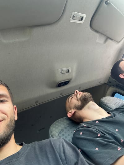 The image shows the interior of a vehicle, likely a van, from a low angle looking upwards at the roof lining. Two men are visible. On the left, a portion of a man's face is visible, with his eye, nose, and beard in focus. He appears to be looking forward or slightly to the left. In the center and right of the frame, a second man is lying down, his head resting on a seat, with his eyes closed and his face tilted upwards. He has a beard and is wearing a dark t-shirt with a pattern of pink flamingos. The vehicle's ceiling has a sun visor on the right, and a light fixture and ventilation controls are visible on the ceiling. The overall environment is dim and appears to be daytime. There is no indication of the city of Tulovo, Bulgaria, within the image itself.