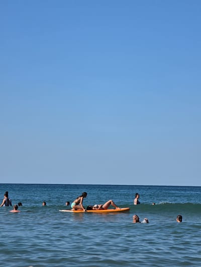 A group of people are in the sea near Ahtopol, Bulgaria, on a clear, sunny day. The water is blue with gentle waves. In the foreground, several individuals are swimming, with their heads visible above the water. Further out, two women are on an orange inflatable surfboard. One woman is kneeling on the board, and the other is lying down. More people are swimming and wading in the background. The sky is a clear, pale blue.