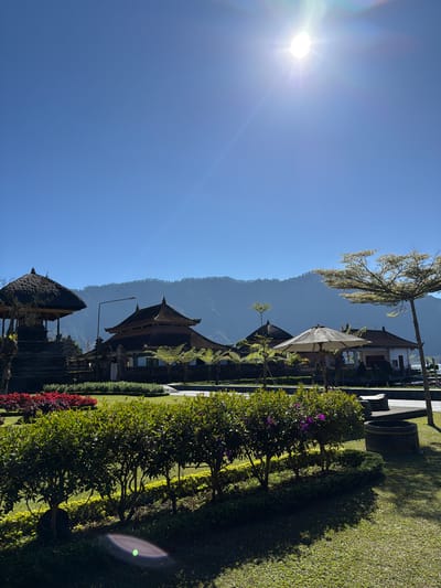 The image captures a view of Pura Ulun Danu Bratan, a water temple complex located in Baturiti, Bali, Indonesia, on a bright, sunny day. The sky is a clear, vibrant blue with the sun prominently positioned in the upper right quadrant, casting a bright glare and lens flare. In the foreground, a meticulously maintained garden features neatly trimmed green bushes and a strip of red flowers. Beyond the garden, several traditional Balinese structures with tiered, dark-tiled roofs are visible. One structure on the left has a thatched roof. A large white umbrella provides shade near one of the buildings. In the background, hazy, verdant mountains rise against the horizon. The overall scene conveys a sense of tranquility and cultural beauty.