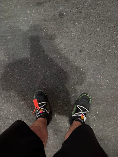 The image displays a top-down view of a person's lower legs and feet standing on a dark, textured paved surface. The ground is a speckled, dark gray material, likely asphalt or coarse concrete, with numerous small light and dark aggregates visible.

The person is wearing dark, possibly black, trousers or shorts that cover the upper legs, and black athletic sneakers. A prominent feature of the sneakers is their bright white laces, which stand out against the dark shoe upper. Both shoes have a distinctive neon green, spiky outsole, creating a strong color contrast.

Attached to the laces of the right shoe is a noticeable orange plastic tag, characteristic of the Off-White brand. White text, partially visible on the tag, indicates Off-White branding. Additionally, a small, multicolored charm or accessory, primarily pink and yellow, is clipped onto the laces of the right shoe.

The lighting in the scene appears dim, casting a faint, diffuse shadow from the left leg onto the pavement. This suggests the image was taken during evening, night, or in an environment with subdued artificial lighting. There are no visible cues regarding weather conditions. The setting is an urban or outdoor paved area, consistent with the context of Ihtiman, Bulgaria. The image captures the person in a moment of standing or walking.