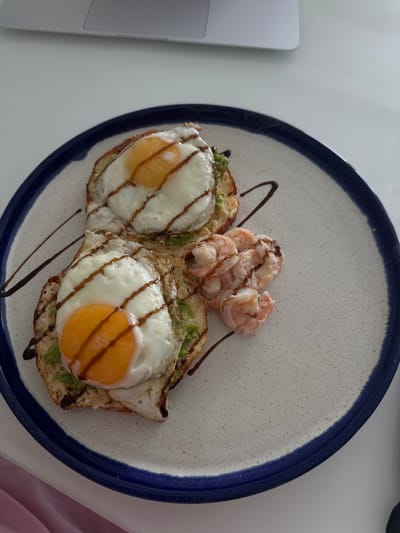 Two pieces of toast topped with mashed avocado and fried eggs are presented on a plate. The eggs have runny yolks and are drizzled with a dark sauce. A small pile of cooked shrimp sits to the right of the toast. The plate has a speckled white surface with a dark blue rim. The setting appears to be indoors, with a portion of a laptop visible in the upper portion of the image. There is no information in the image to suggest a location in Ivano-Frankivsk, Ukraine.