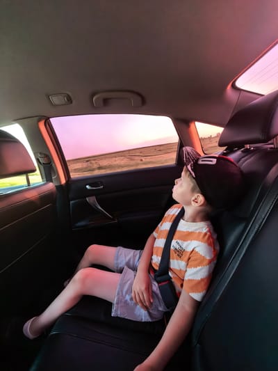 The image shows a young boy sitting in the back seat of a car, looking out the window. The boy is wearing an orange and white striped t-shirt and light-colored shorts. He has a black baseball cap on his head. The car appears to be moving, as indicated by the blurred scenery outside the window, which depicts a rural landscape at sunset with hues of pink and orange in the sky. The interior of the car is dark, with black leather seats. The boy is wearing a seatbelt. The scene is set in Marshanovka, Russia, during the evening.