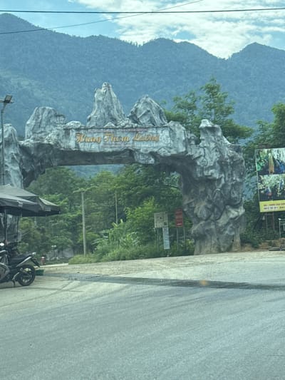 The image shows the entrance to Hang Thúm Lũng, located in Minh Tân Commune, Vietnam. The entrance is a large stone archway with the name of the location written on it. Behind the archway are trees and mountains. In the foreground, there is a road and several parked motorcycles. There are several signs located around the location, partially obscured by the surrounding foliage. The sky is cloudy.