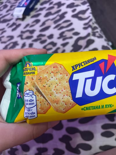 The image shows a close-up of a hand holding a package of TUC crackers. The crackers appear to be flavored with "sour cream and onion" in Russian. The packaging is yellow with blue and red accents. There is text on the packaging that says "ХРУСТАЮЩИЙ TUC" which translates to "CRUNCHY TUC". There is also nutritional information displayed in a small white box, showing that 100 grams of the crackers contain 495 calories and 2074 kJ. The background of the image is a blurry leopard-print fabric. The specific location in Vaganovo, Russia, cannot be determined from this image.