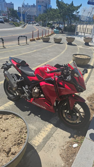 A red Honda motorcycle with an Akrapovic exhaust system is parked on a city street in Ulsan, South Korea. The motorcycle is situated between decorative concrete planters and a yellow line marking the road. In the background, there is a street with traffic, sidewalks, buildings, and a Ferris wheel visible in the distance. The sky is clear.