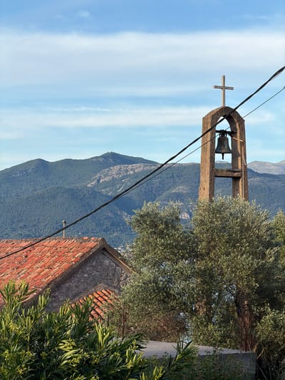 The image depicts a bell tower with a cross on top, situated in the city of Krašići, Montenegro. The tower is made of stone and houses a bell within its structure. In the background, a mountainous landscape is visible under a partly cloudy sky. An olive tree and other foliage partially obscure the view of a red-tiled roof of a building near the bell tower. Electrical wires run across the foreground of the image, slightly obstructing the view of the bell tower and the landscape beyond.