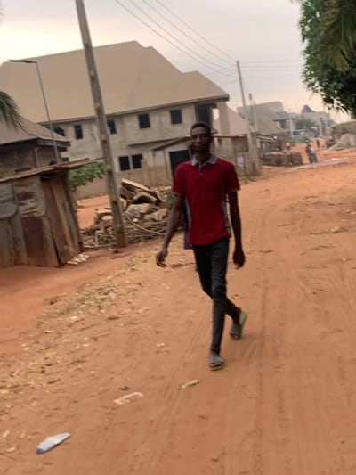 A man stands on a reddish dirt road with construction in the background. He wears a red shirt, dark pants, and green sandals. Litter, including a white plastic bag, is scattered on the ground. There's a wooden structure nearby. A building under construction with visible window openings is in the mid-ground. Utility poles and wires are visible in the distance. The sky is overcast.