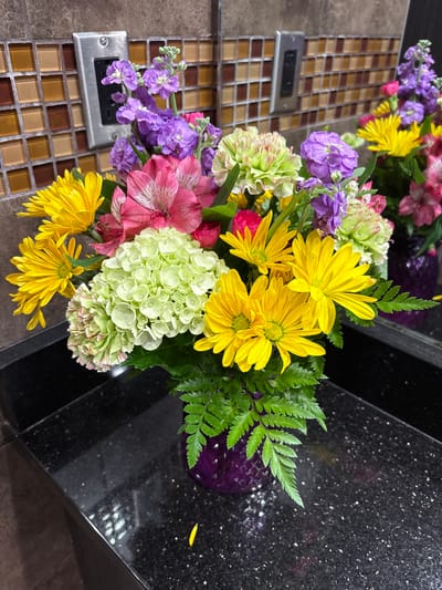 A brightly colored floral arrangement in a purple vase sits on a black, speckled countertop. The bouquet features yellow chrysanthemums, pink alstroemeria, light green hydrangeas, and purple stock flowers. Behind the flowers, a wall with a tiled backsplash in shades of brown and tan is visible. A silver electrical outlet is mounted on the tiled wall. The scene appears to be an indoor setting, possibly a bathroom or kitchen counter.