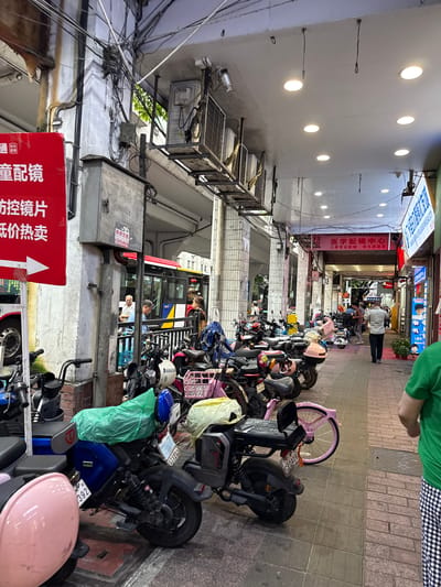 This is a street scene in Yuexiu District, China, featuring a covered walkway lined with parked electric scooters and motorcycles. The walkway is illuminated by overhead fluorescent lights. On the left side, a red sign with white text, possibly an advertisement, is partially visible. Behind the sign, a bus is stopped at what appears to be a bus stop. Several individuals are present, some standing near the bus and others walking along the walkway. The ground is paved with brick-like tiles. Overhead, electrical wires are densely strung across the ceiling of the walkway. The atmosphere is urban, with a mix of commercial activity and transportation infrastructure.