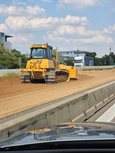 The image shows a yellow bulldozer with the text "DBS Schmitt" on its cab, operating on a dirt road adjacent to a concrete barrier. The bulldozer is positioned on the right side of the frame, with its tracks partially embedded in the sandy soil. The cab of the bulldozer is visible, featuring windows and what appears to be an orange warning light on top. In the background, there are trees and buildings, suggesting an urban or suburban setting. A white truck with a blue trailer is also visible further down the road, partially obscured by the bulldozer. The sky is bright blue with scattered white clouds, indicating it is daytime and likely sunny. The concrete barrier next to the bulldozer has yellow markers embedded in it at intervals. A reflection of the yellow bulldozer and surrounding scene is visible in a shiny surface to the right of the image, possibly a vehicle mirror. The image was taken in Rüsselsheim am Main, Germany. The scene appears to be a construction site, possibly related to roadwork or infrastructure development, given the presence of the bulldozer and the road surface. There are no visible people in the image. The overall impression is one of ongoing construction activity on a clear day.