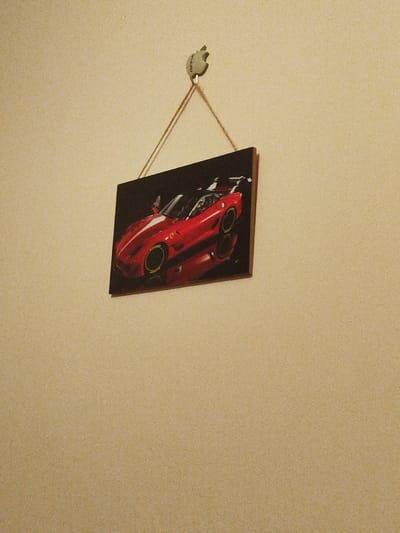 The image shows a red Ferrari sports car hanging on a wall in a room. The car is depicted in a framed picture on a beige wall. The picture is hanging on a string and a small, green decorative hook. The picture is mounted on a beige wall. The image is located in Gitaru, Kenya (latitude: -1.232, longitude: 36.676). There are no people in the scene, no actions, interactions, or other objects except for the picture and the hook on the wall. The room is lit from above and the walls are beige. The picture is in focus, while the wall behind it is slightly blurred.
