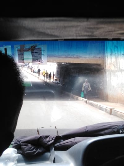The image presents a scene from inside a vehicle, looking out through the front windshield in Kikuyu Township ward, Kenya. The foreground shows the vehicle's dark dashboard, with a crumpled black fabric item (possibly a jacket or bag) displaying a "NY" logo on the right side. The left side of the frame is occupied by the back of a person's head, indicating a driver or front passenger.

Affixed to the upper center of the windshield are two transparent stickers, one prominently displaying "ILADE OIL," a Kenyan telephone number starting with "+254," and "KRA PIN."

Beyond the windshield, a paved road stretches forward, passing under a concrete bridge or overpass. The underside of the bridge is shadowed and appears dark, while the sky above is bright blue. On the left side of the road, a group of approximately six to eight pedestrians, consisting of men and women in casual attire, walk away from the viewer, heading towards or through the underpass. On the right side, a solitary individual, dressed in light-colored clothing, walks on a raised concrete sidewalk, carrying a green bag in their left hand, also moving away from the viewer and approaching the shaded area beneath the underpass. The concrete supporting wall of the underpass on the right shows some discoloration and faint markings. The scene depicts pedestrian movement alongside a road infrastructure in a daylight setting.