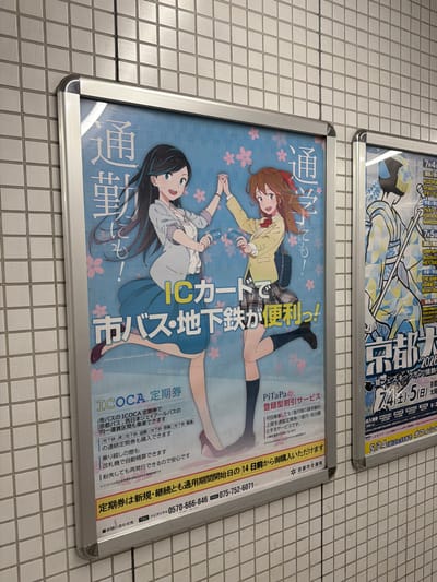 The image shows a poster on a tiled wall, likely in a subway station or public transport area in Kyoto, Japan. The poster is for ICOCA and PiTaPa, contactless smart cards used for public transportation. The poster features an anime-style illustration of two young women with pink flowers in the background. One woman has long black hair and is wearing a white blouse and blue skirt, holding an ICOCA card. The other woman has brown hair with a red ribbon and is wearing a yellow blazer and plaid skirt, also holding a card. Text on the poster promotes the convenience of using these cards for city buses and subways. Another poster to the right is partially visible and appears to be advertising an event in Kyoto, with text including "Kyoto Da..." and dates. The overall impression is a promotional display for public transportation services in Kyoto.